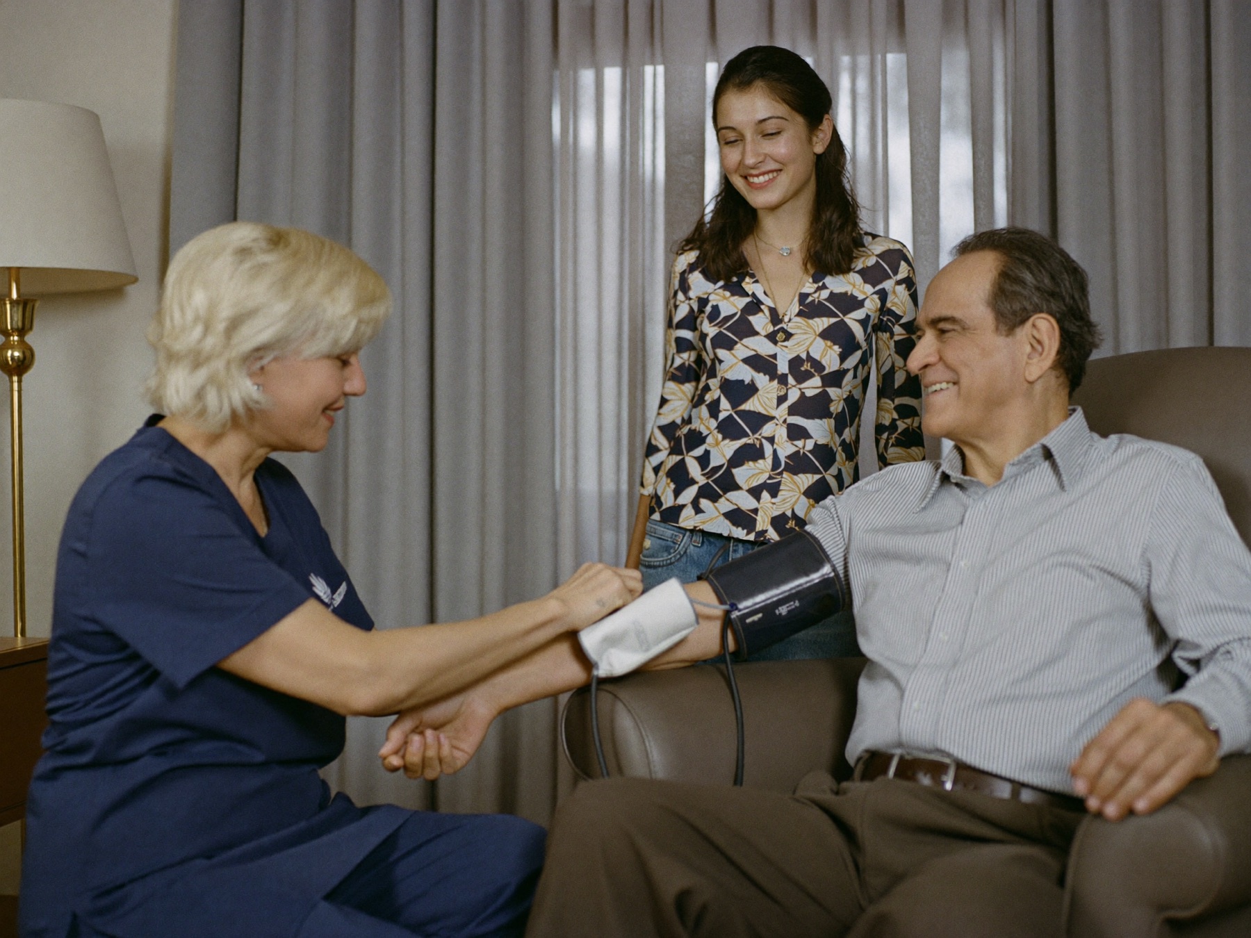 A nurse checking an elderly man's blood pressure at home, his daughter beside him